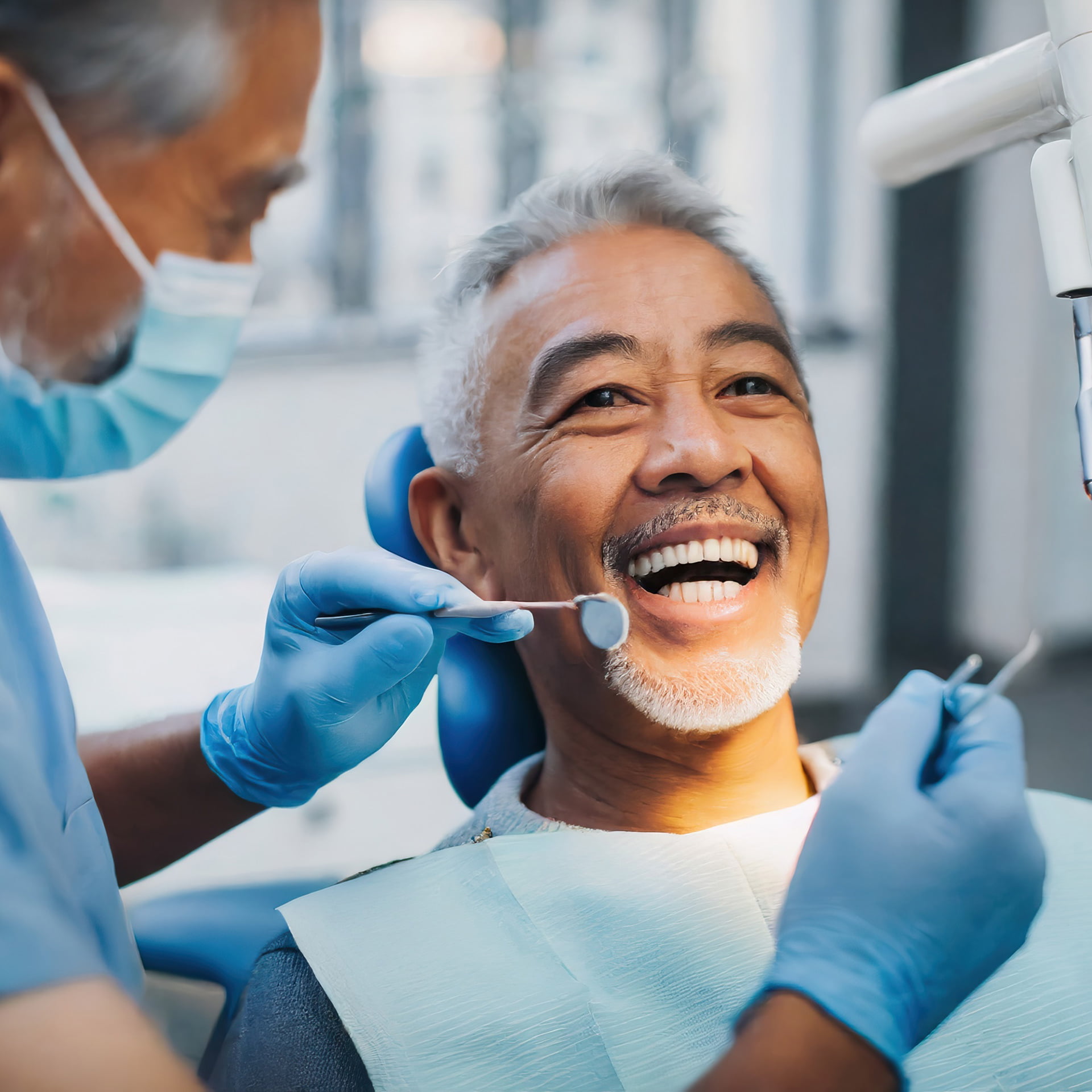 A smiling woman with brown hair reclining comfortably in a dentist's chair, exhibiting a relaxed and positive demeanor. She's wearing a casual olive green jacket and the chair is upholstered in blue, indicating a clean and professional dental care environment.