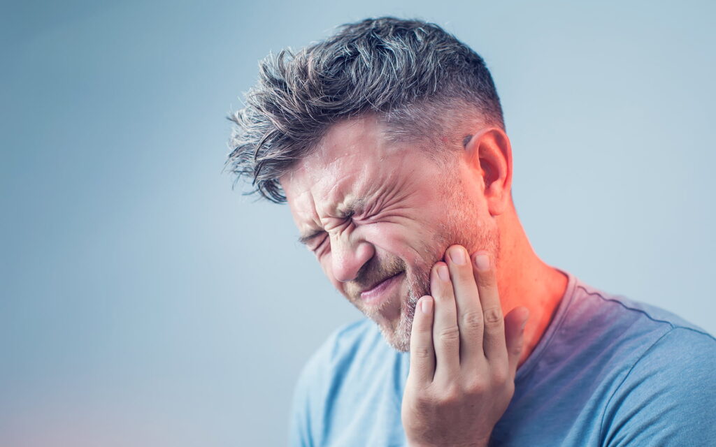 A middle-aged man with graying hair, squinting and grimacing in pain, holds his cheek and jawline suggesting a toothache or jaw discomfort. He wears a casual blue t-shirt, set against a soft blue gradient background.