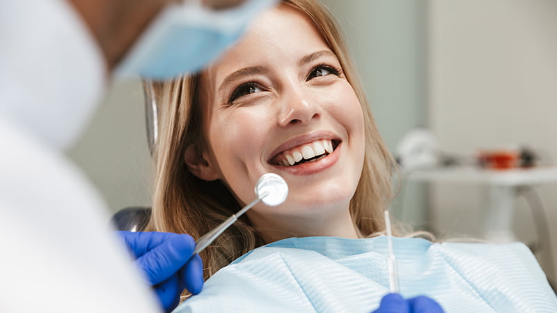 A cheerful female patient receiving a dental check-up is smiling at her dentist. She is wearing a protective bib while the dentist, in white attire and gloves, holds dental tools. The close-up captures the positive interaction and the detail of the dental instruments, suggesting a comfortable and friendly dental care experience.