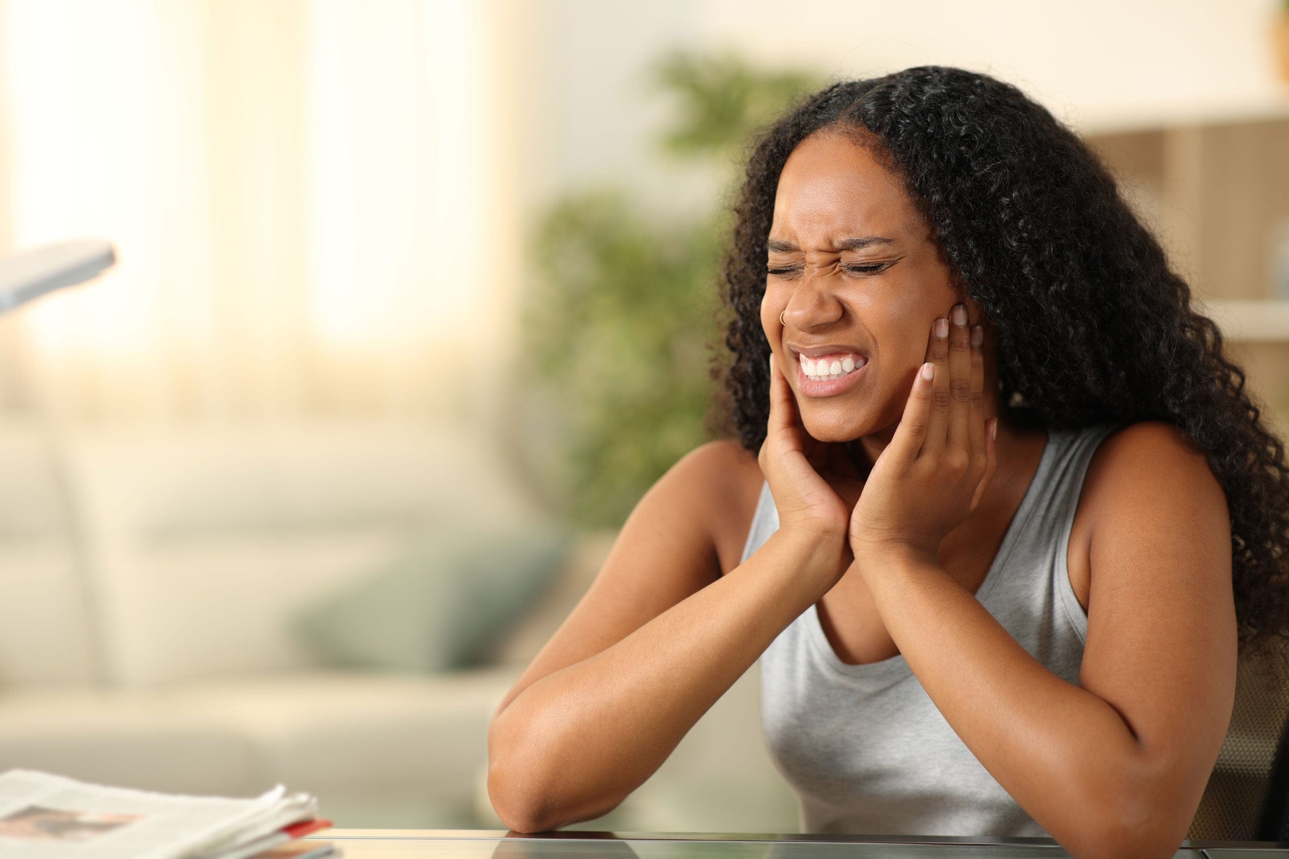 Woman iwearing a gray tanktop and sitting in her living room is holding her jaw as she is experiencing dental pain.
