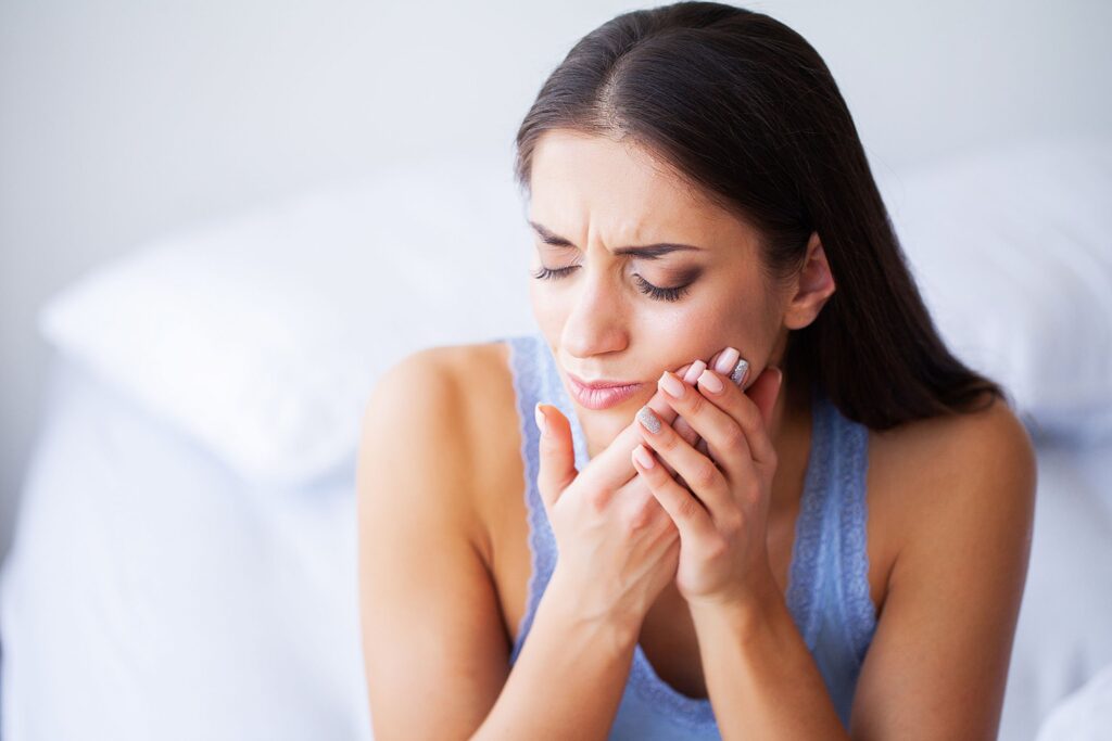 A concerned woman sitting on a bed, holding her jaw and cheek, with a pained expression that suggests a toothache, in a bedroom setting with a white pillow in the background.