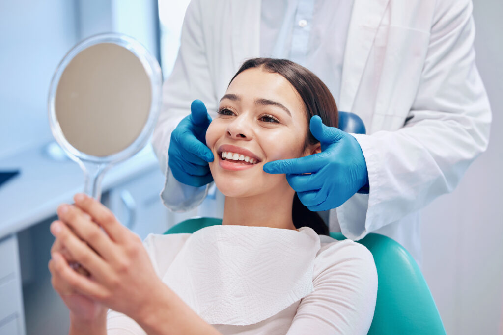 Patient smiling in dental chair while dentist with gloves checks teeth and patient holds a mirror.