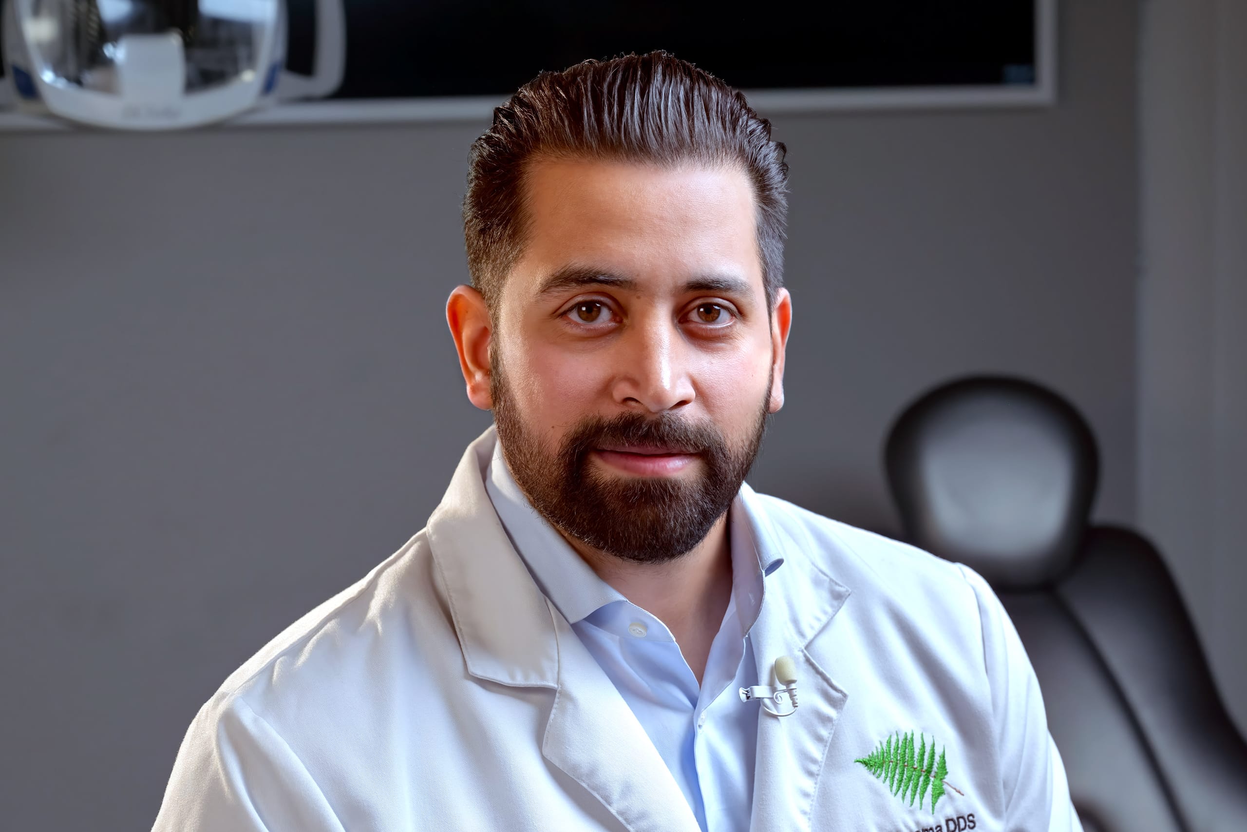 An image of Dr. Cheemsa sitting in a white lab coast. Behind him is a dental chair and light indicating he is in a patient treatment room.