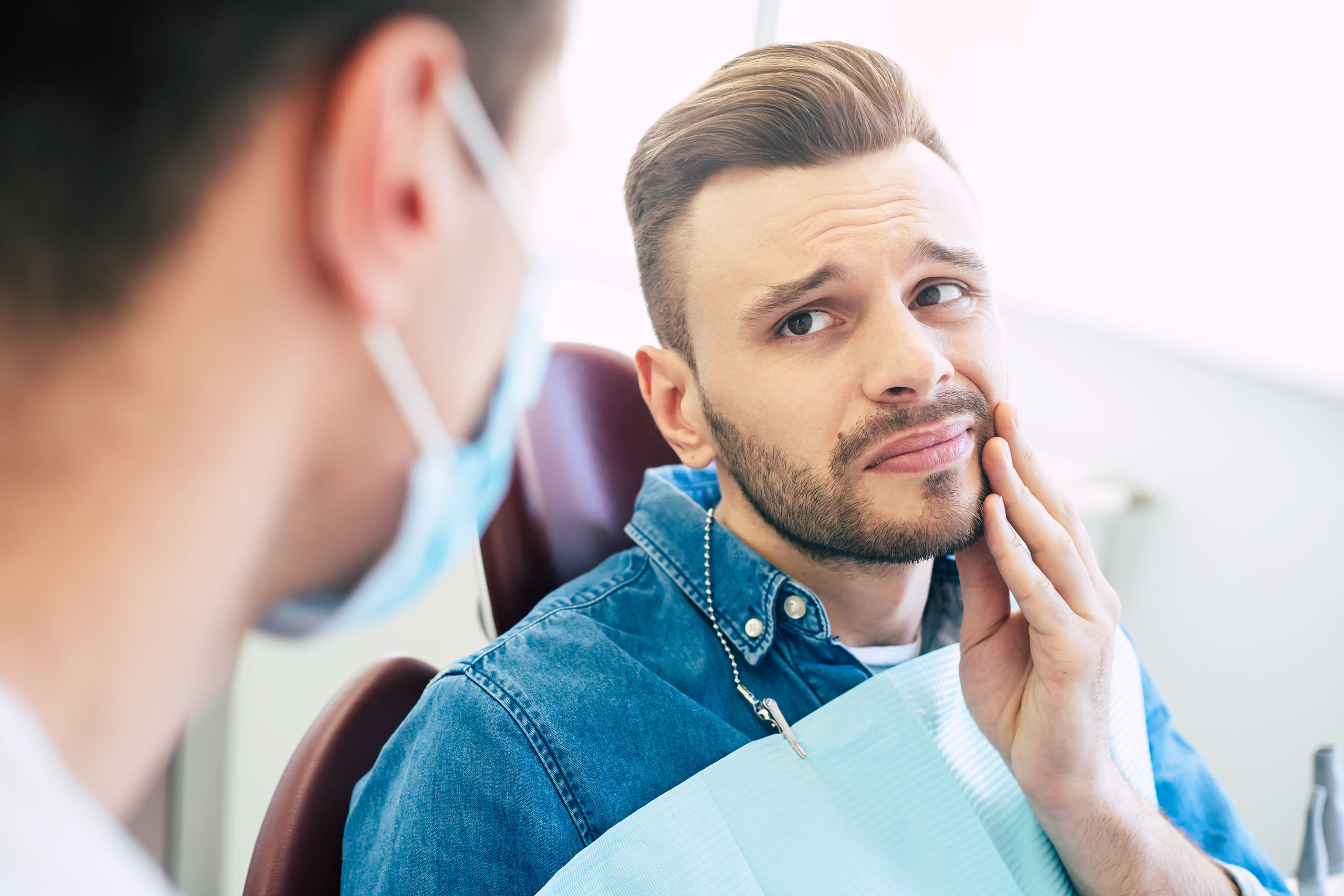 Adult patient in a dental chair touching his cheek while discussing tooth pain with a masked dentist.