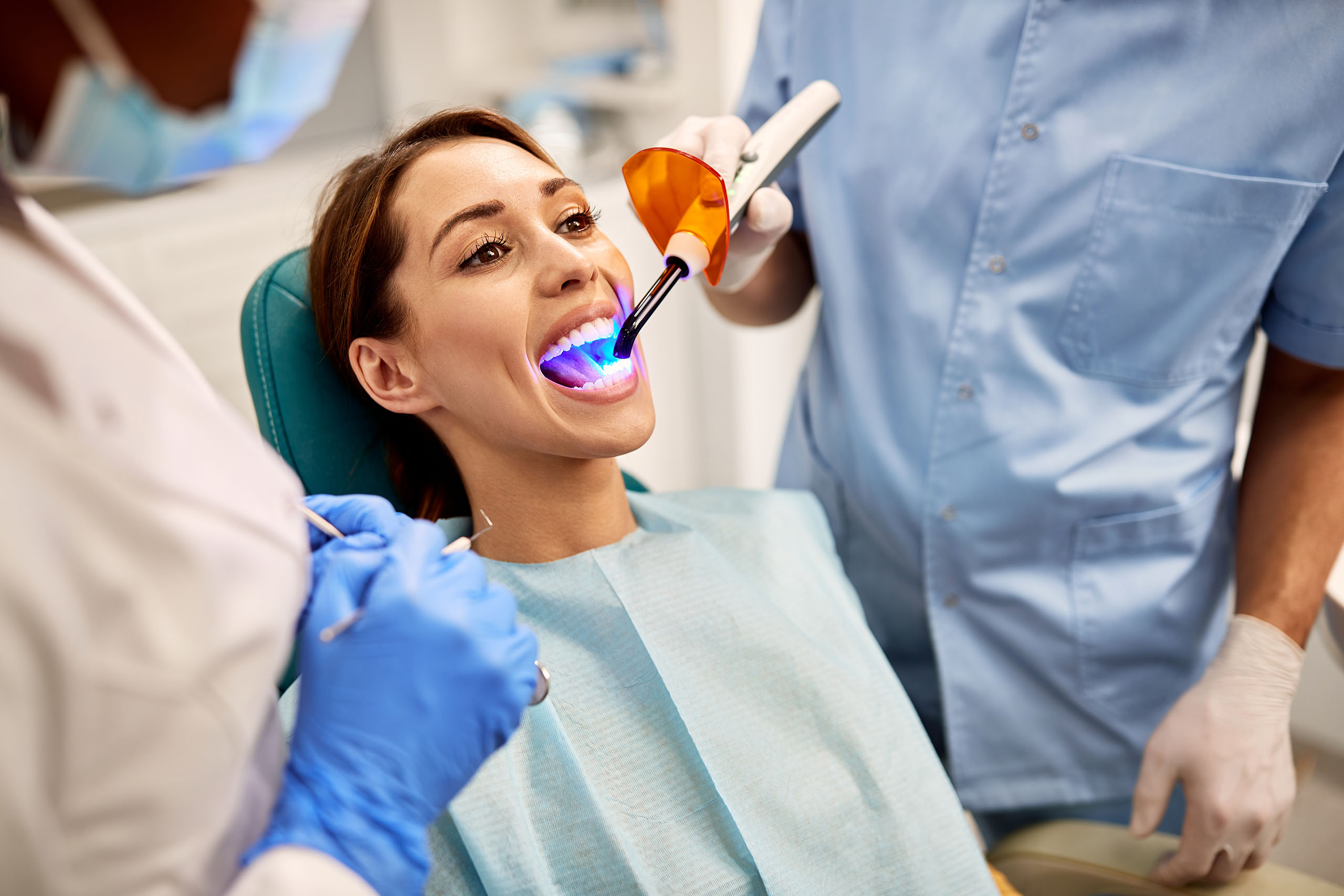 Dentist using a blue curing light to harden a dental filling while a patient reclines in a treatment chair.