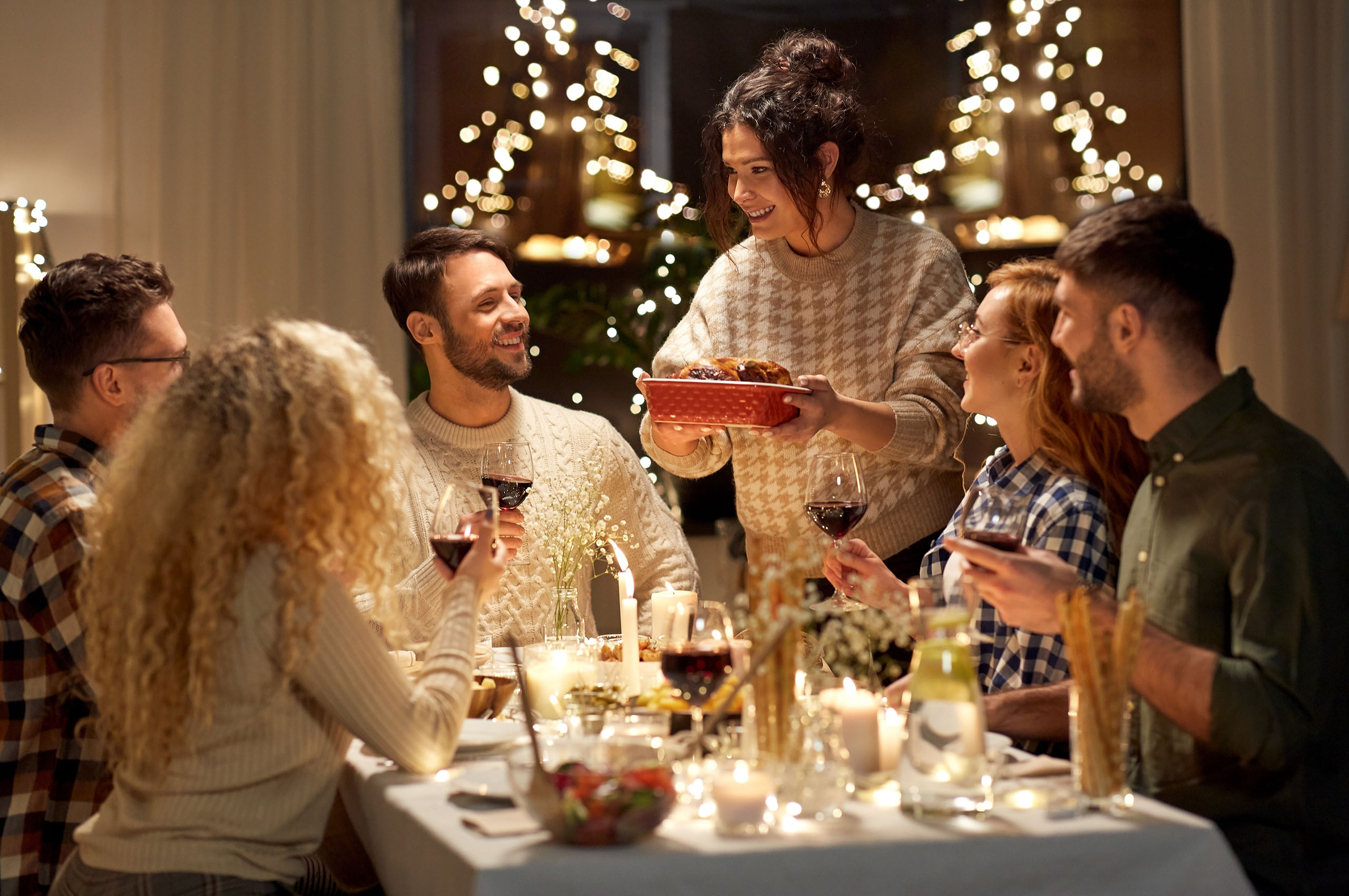 Warm holiday dinner gathering with six friends smiling and sharing a freshly served dish at a candlelit table.