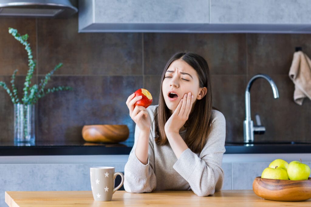 Woman holding an apple and touching her cheek, showing discomfort while eating at a kitchen table.
