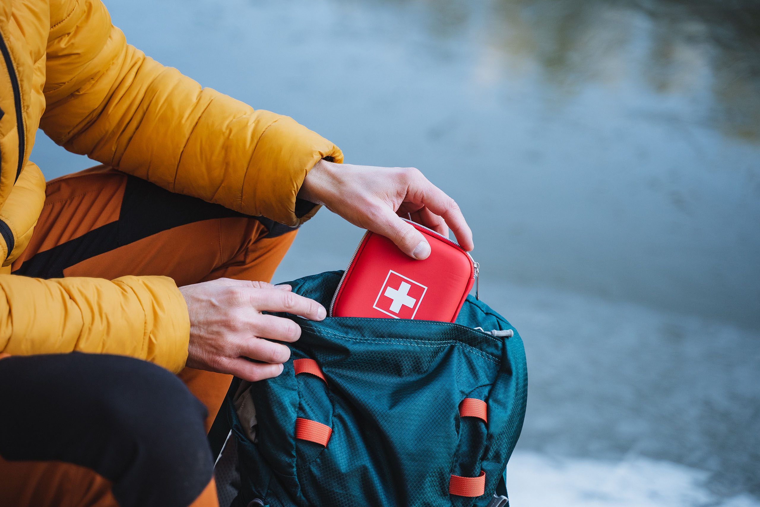 Person placing a red first aid kit into a backpack outdoors near a body of water.