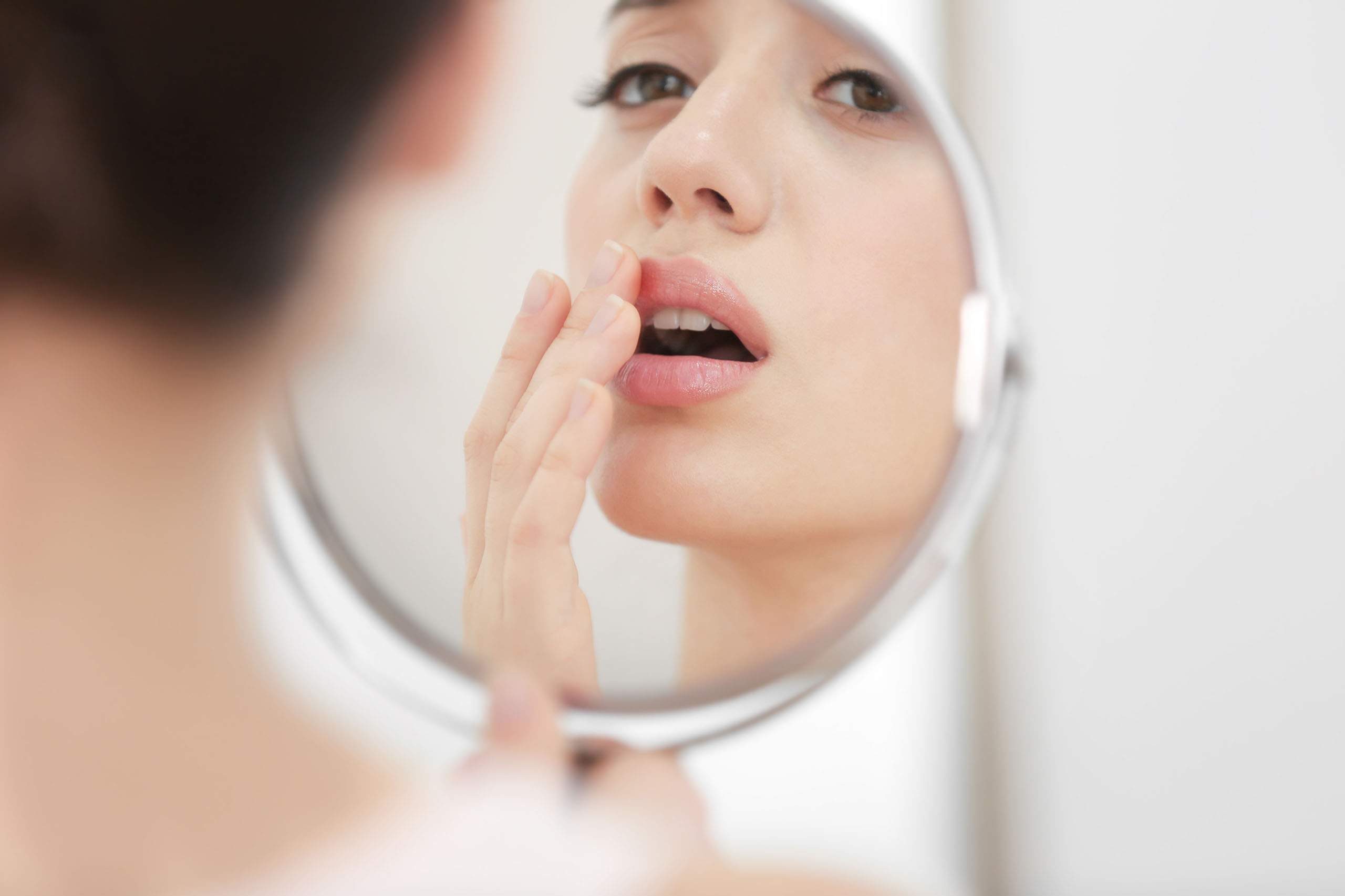 Person examining a cold sore on their lip in a handheld mirror, touching the affected area to check irritation