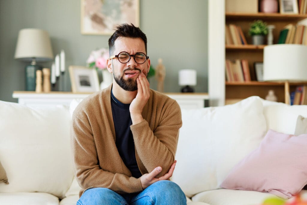 Man sitting on a couch holding his jaw in pain, suggesting toothache or dental discomfort at home
