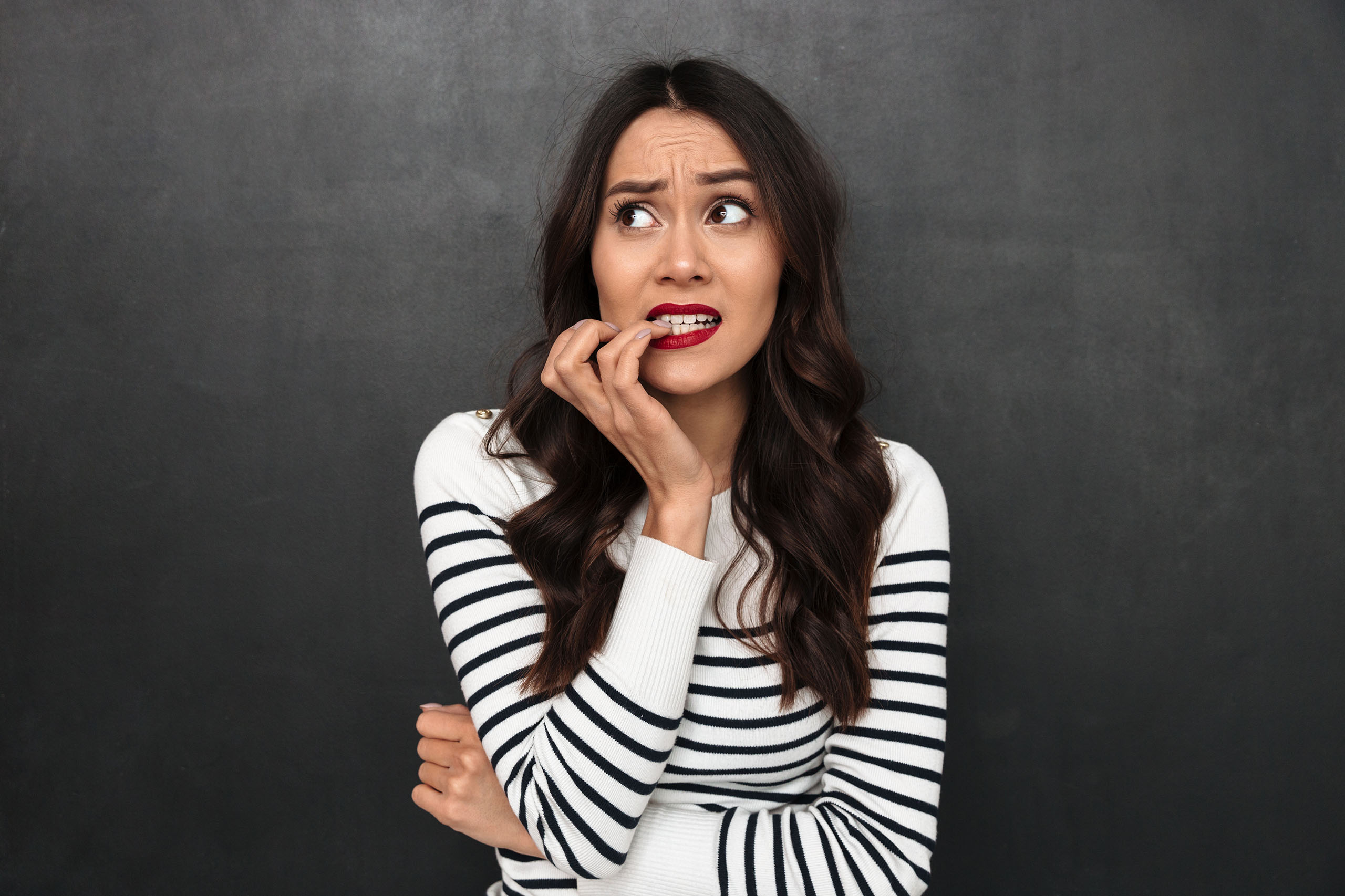 Anxious woman biting her fingernails, looking up with worried expression against a dark background