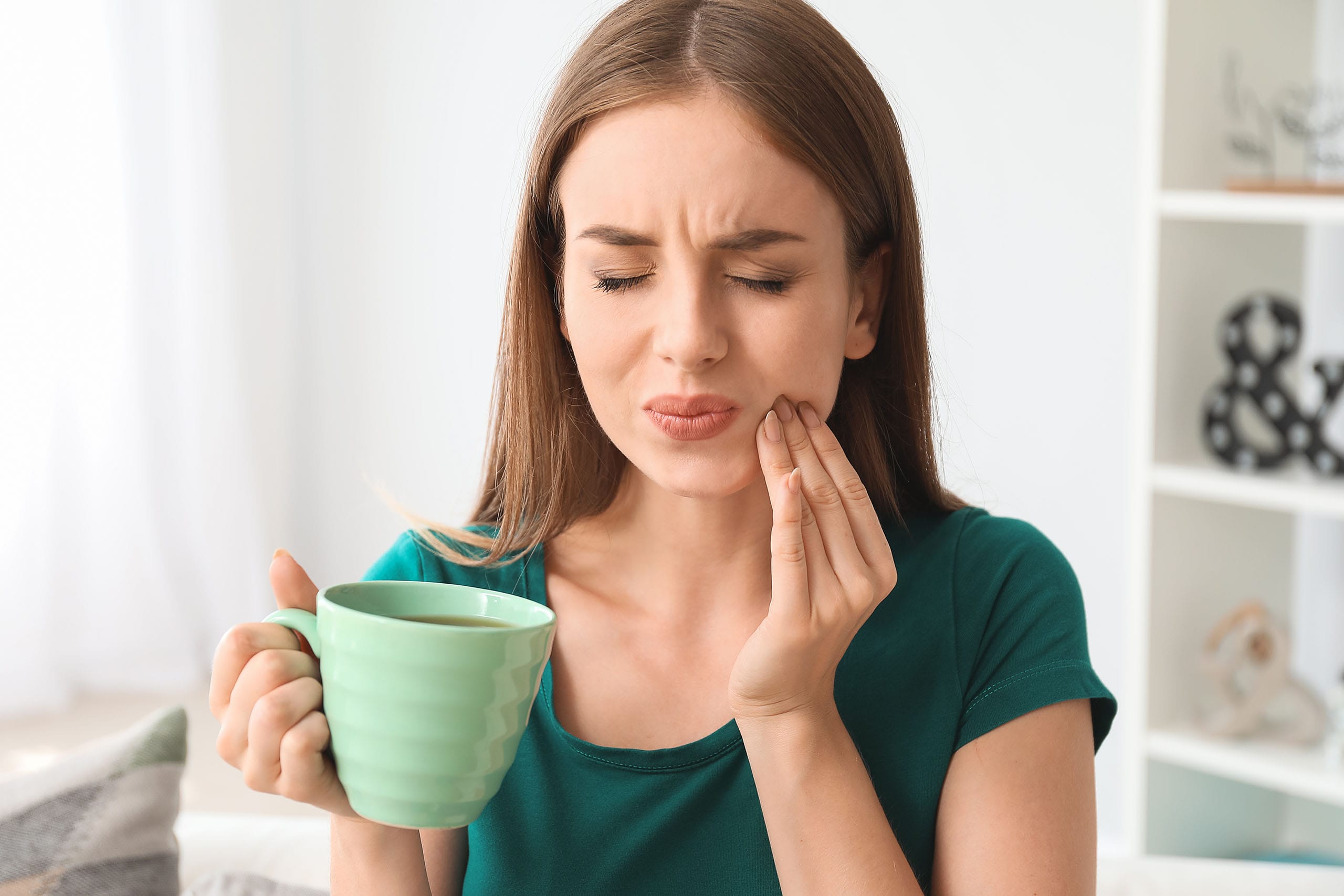 Woman holding a mug, pressing her cheek with eyes closed, showing discomfort from tooth pain at home